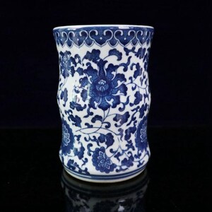 May include: A cylindrical white and blue ceramic vase with a floral pattern. The vase features a repeating blue floral design on a white background. The top rim has a decorative blue border. The vase is likely used for decorative purposes.