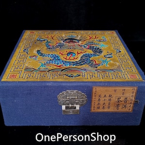 Op de afbeelding: Een blauwe fluwelen doos met een gouden deksel met een ontwerp van een blauwe en rode draak. Het deksel is versierd met een gouden rand en een herhalend patroon. De doos heeft een metalen sluiting en een label met Chinese karakters.