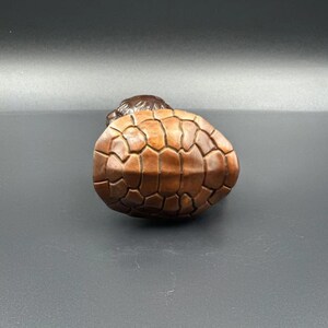 以下が含まれることがあります： 茶色とベージュの甲羅を持つ木製の亀の置物。亀は背中に仰向けになっており、手足と頭を引っ込めている。