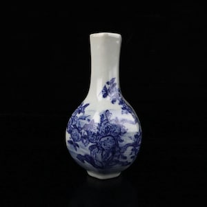 Puede incluir: Un jarrón de cerámica blanco y azul con un cuerpo bulboso y un cuello estrecho. El jarrón presenta un diseño floral pintado a mano en tonos azules. El diseño incluye flores y follaje. El jarrón es un objeto decorativo.