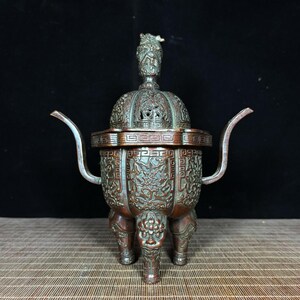 May include: An antique bronze incense burner with intricate dragon and geometric patterns. The burner has a domed lid with a decorative finial, curved handles, and three ornate legs. The overall color is a mix of bronze and verdigris.