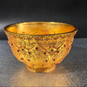 May include: An ornate gold-coloured bowl with intricate embossed patterns and embedded red and green gemstones. The bowl features a detailed design around the exterior, with a decorative base. Likely a decorative or ceremonial item.