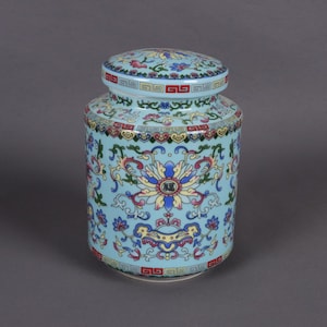 May include: A light blue ceramic jar with a lid, adorned with a floral pattern in shades of pink, yellow, and blue. The jar features a repeating geometric border and a central floral design with the Chinese character for "blessing".