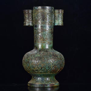 Puede incluir: Un jarrón cilíndrico de bronce con una pátina verde desgastada y acentos de color marrón oscuro. La superficie presenta intrincadas tallas en relieve de figuras y patrones en el cuello y el cuerpo bulboso, con dos pequeñas asas tubulares.