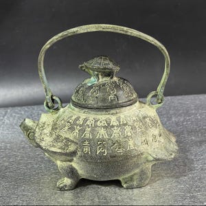 May include: Antique bronze teapot shaped like a turtle, with a handle and a lid. The teapot is covered in intricate carvings and Chinese characters. The lid is topped with a small turtle figurine.