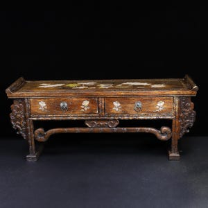 Op de afbeelding: Een kleine, antieke houten kast met twee lades. De kast is bruin met bloemeninleg op de bovenkant en de ladefronten. De zijkanten en poten hebben sierlijke gravures. De ladetrekker zijn klein en donker.