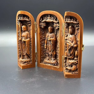 Puede incluir: Un santuario plegable de madera de tres paneles con intrincados tallados que representan tres figuras. El santuario está cerrado y las figuras son visibles a través de los paneles tallados.