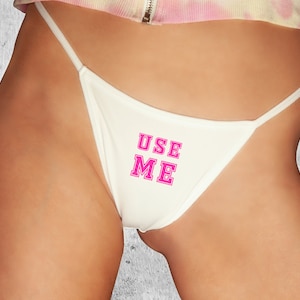 May include: Off-white thong underwear with thin straps. The text "USE ME" is printed in pink, outlined in a darker pink, across the front of the underwear. The garment is made of a smooth fabric.