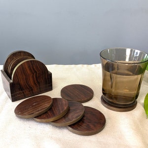 Puede incluir: Juego de seis posavasos de madera marrón con ribete de latón, en un soporte, con un vaso de agua sobre un mantel beige.