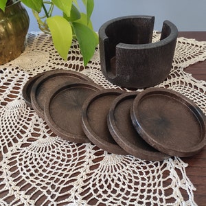 Può includere: Set di sei sottobicchieri in legno marrone scuro con un supporto. I sottobicchieri sono rotondi e hanno una finitura liscia e lucida. Il supporto è anch'esso in legno e ha una forma cilindrica.