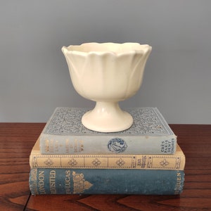 May include: A cream-colored, tulip-shaped pedestal bowl sits atop a stack of three vintage books. The books are in shades of blue and tan, with gold lettering and decorative patterns. The background is a solid gray.