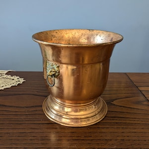 May include: A copper-coloured metal planter with a decorative lion's head and ring handle. The planter has a wide, flared rim and a pedestal base. The surface shows signs of age and use, adding to its vintage appeal.