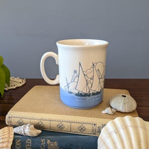 Op de afbeelding: Witte keramische mok met een blauwe basis en een nautisch ontwerp met zeilboten. De mok heeft een gebogen handvat en staat op een stapel boeken. Schelpen en een plant staan op de achtergrond.