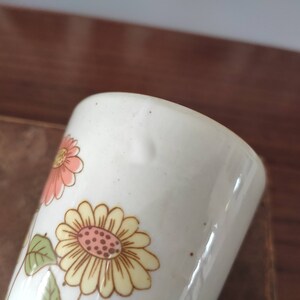 May include: White ceramic mug with a floral design featuring pink, yellow, and brown flowers. The mug has a slightly chipped edge.