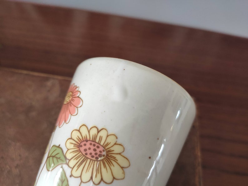 May include: White ceramic mug with a floral design featuring pink, yellow, and brown flowers. The mug has a slightly chipped edge.