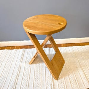 Op de afbeelding: Een lichtgekleurde houten klapstoel met een ronde zitting en een gat. De kruk heeft een X-vormige basis en is gemaakt van lichtgekleurd hout. De kruk staat op een wit tapijt en een houten vloer.