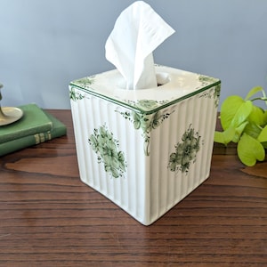 Può includere: Un copriscatola per fazzoletti quadrato in ceramica bianca con dettagli a coste verticali e motivi floreali verdi dipinti a mano su ogni lato e angolo. Un fazzoletto bianco fuoriesce dall'apertura superiore del contenitore.
