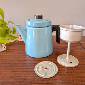 Puede incluir: Una cafetera vintage de esmalte azul claro con asa y tapa negras. La imagen también muestra una cesta de filtro de metal y un disco de metal perforado. Los artículos están colocados sobre una superficie de madera.