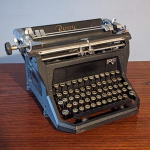 Puede incluir: Una máquina de escribir negra vintage con detalles plateados y un teclado completo. La palabra "Rooy" está inscrita en la parte superior. La máquina de escribir se encuentra sobre una superficie de madera, mostrando su diseño clásico y detalles mecánicos.