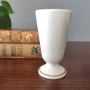 May include: A white ceramic drinking glass with a speckled glaze and a brown rim. The glass has a pedestal base.