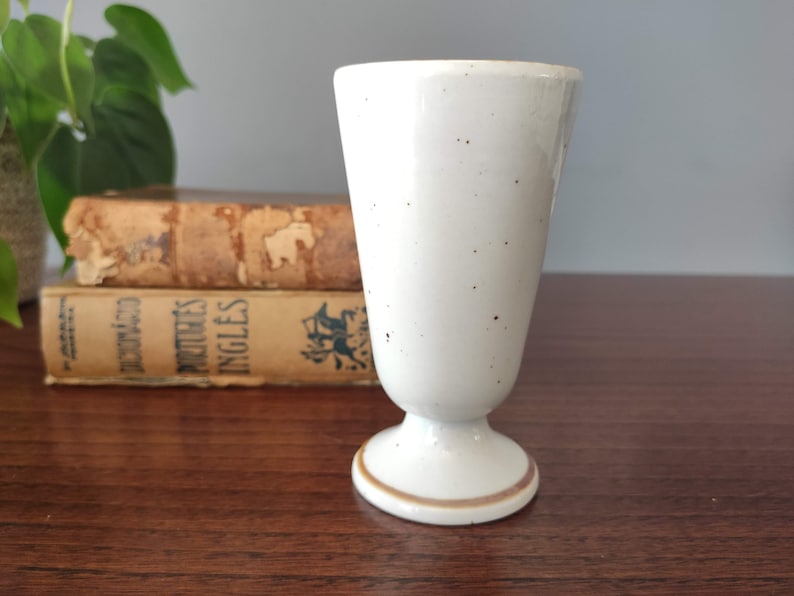 May include: A white ceramic drinking glass with a speckled glaze and a brown rim. The glass has a pedestal base.