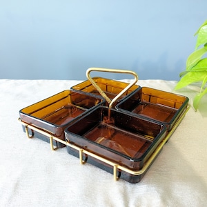 Puede incluir: Un plato de servir de vidrio ámbar de cuatro secciones con un marco y un asa de metal dorado. El plato está sobre una superficie blanca.