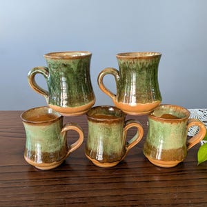 Könnte beinhalten: Fünf handgefertigte Keramikbecher mit rustikalem, glasiertem Finish. Die Becher haben ein grün-braunes Farbschema mit einem beigen Boden und Griff. Die Becher sind auf einer dunklen Holzoberfläche angeordnet.