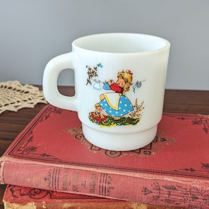 May include: White ceramic mug with a colorful illustration of a girl hanging laundry on a clothesline. The girl is wearing a blue and white polka dot dress and a red shirt. The mug is sitting on top of a red book.