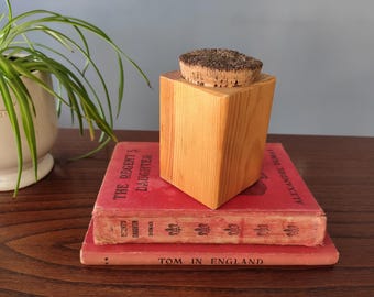 Op de afbeelding: Een houten, vierkante container met een kurken deksel staat op twee vintage rode boeken. De container is gemaakt van lichtgekleurd hout. De boeken hebben gouden letters en zijn op elkaar gestapeld.