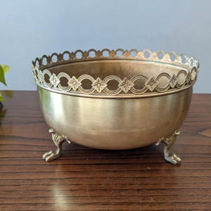 Può includere: Una ciotola in metallo color oro con un bordo traforato decorativo con motivi floreali e ovali. La ciotola poggia su tre piccoli piedini a forma di artiglio. La ciotola è probabilmente utilizzata per scopi decorativi.