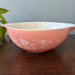 May include: A vintage pink mixing bowl with a white interior and a decorative white floral pattern. The bowl has a handle-like spout on one side. A classic kitchenware item.