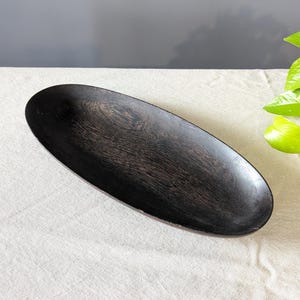 Pode incluir: Uma bandeja de madeira de formato oval alongado com um acabamento em castanho escuro profundo. A superfície da bandeja apresenta um padrão visível de veios de madeira natural, com um interior liso e côncavo e uma borda curva e rasa.
