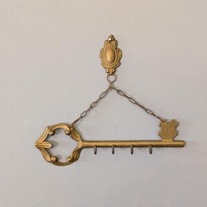 May include: A gold-colored key-shaped wall-mounted key holder. The key has an ornate design with a decorative top and a series of small hooks for hanging keys. It is suspended by a chain from a decorative wall mount.