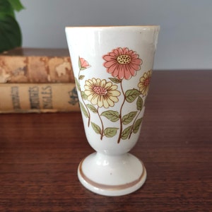 May include: A white ceramic vase with a footed base and a floral pattern. The vase features pink and yellow flowers with green leaves.