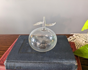 Könnte beinhalten: Ein klarer Glasapfel mit Stiel und Blättern. Der Apfel liegt auf einem Stapel Bücher.
