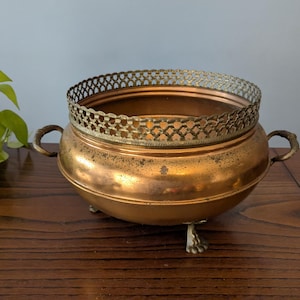 Può includere: Una ciotola vintage in rame con bordo e manici decorativi in ottone. La ciotola poggia su tre piedini ornati. Il rame ha una calda patina invecchiata, che suggerisce la sua natura antica. Un pezzo decorativo per l'arredamento della casa.