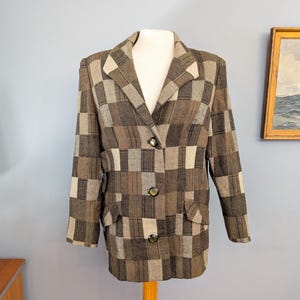 Puede incluir: Una chaqueta de manga larga con un patrón de cuadros en tonos marrón, beige y crema. La prenda cuenta con solapas de muesca, tres botones oscuros en el cierre frontal y dos bolsillos con solapa en la cintura. La tela tiene textura tejida.