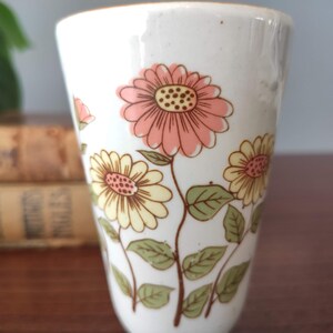 May include: A white ceramic mug with a floral design. The flowers are pink, yellow, and green. The mug is a tall, slender shape.