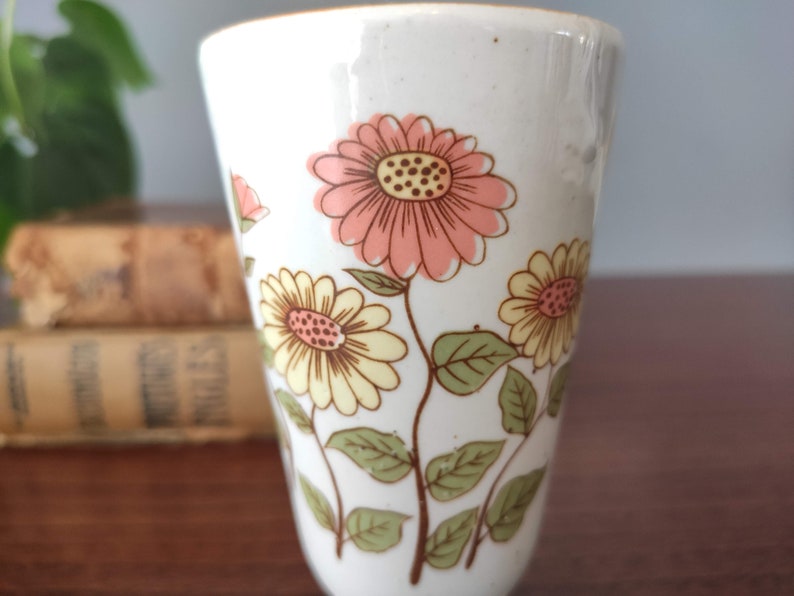 May include: A white ceramic mug with a floral design. The flowers are pink, yellow, and green. The mug is a tall, slender shape.