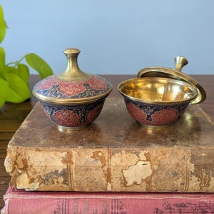 May include: Two small, decorative brass bowls with lids. One bowl is open, revealing a gold interior. The bowls have a red and blue floral design. They sit on a stack of antique books with worn covers.