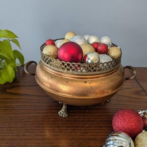 Pode incluir: Uma tigela cor de cobre com uma borda decorativa em filigrana, cheia de enfeites de Natal vermelhos, dourados e prateados. A tigela assenta em três pés ornamentados e tem duas pegas. Enfeites adicionais estão espalhados por perto.