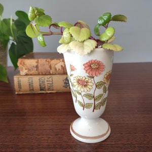 May include: A white ceramic vase with a floral pattern featuring pink, yellow, and green flowers. The vase has a flared top and a pedestal base. A small green plant is in the vase.