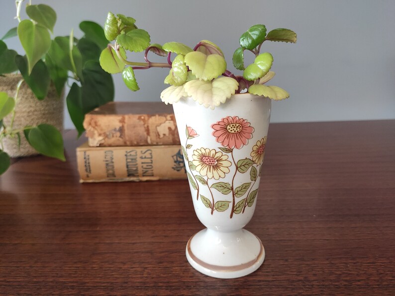 May include: A white ceramic vase with a floral pattern featuring pink, yellow, and green flowers. The vase has a flared top and a pedestal base. A small green plant is in the vase.