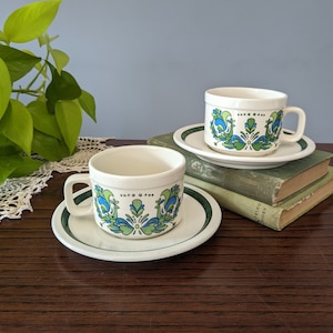 May include: Two white ceramic teacups with green and blue floral designs and matching saucers. The cups and saucers have a green band around the rim.