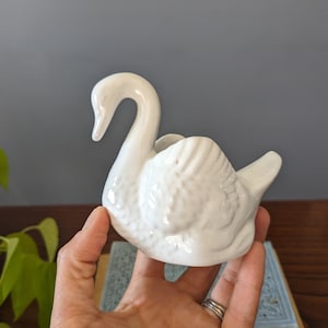 May include: White ceramic swan figurine with a detailed design. The swan has a long neck and a curved body. It is a decorative piece for a home or garden.