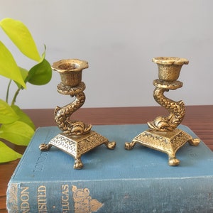 May include: Two gold metal candle holders shaped like dolphins with ornate details. The candle holders are sitting on a blue book.