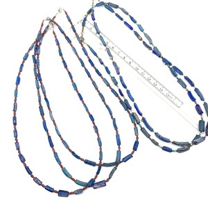 Peut inclure: Trois colliers de perles de verre bleu et rouge. Les colliers sont faits de petites perles cylindriques. Les perles sont d'un bleu fonc&eacute; et d'un brun rouge&acirc;tre. Les colliers sont dispos&eacute;s sur une surface blanche avec une r&egrave;gle pour l'&eacute;chelle.