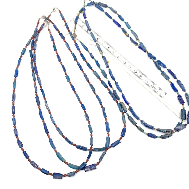 Peut inclure: Trois colliers de perles de verre bleu et rouge. Les colliers sont faits de petites perles cylindriques. Les perles sont d'un bleu fonc&eacute; et d'un brun rouge&acirc;tre. Les colliers sont dispos&eacute;s sur une surface blanche avec une r&egrave;gle pour l'&eacute;chelle.