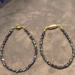 Op de afbeelding: Twee armbanden bestaande uit kleine, donkere, onregelmatige, metaalachtig ogende kralen. De linkerarmband heeft een goudkleurige sluiting, terwijl de rechterarmband is voorzien van een cilindervormige goudkleurige magnetische sluiting.