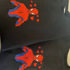 Puede incluir: Dos sudaderas negras con una imagen bordada de Spiderman con corazones a su alrededor, en rojo y azul.